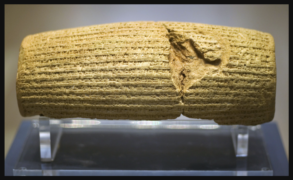 Front view of a barrel-shaped clay cylinder resting on a stand. The cylinder is covered with lines of cuneiform text. British Museum.